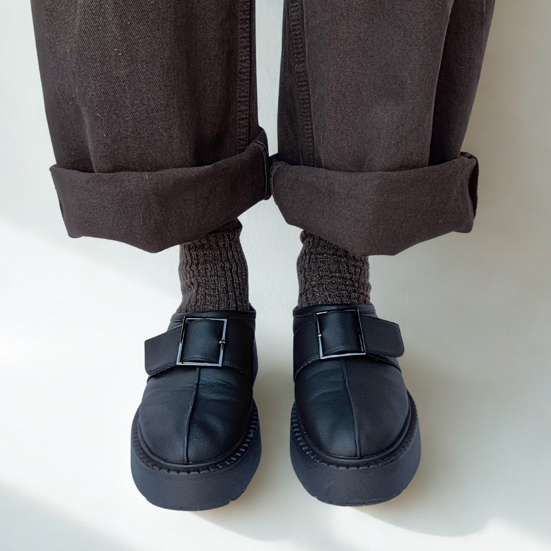 Person wearing dark brown pants, gray socks, and black shoes on a white background
