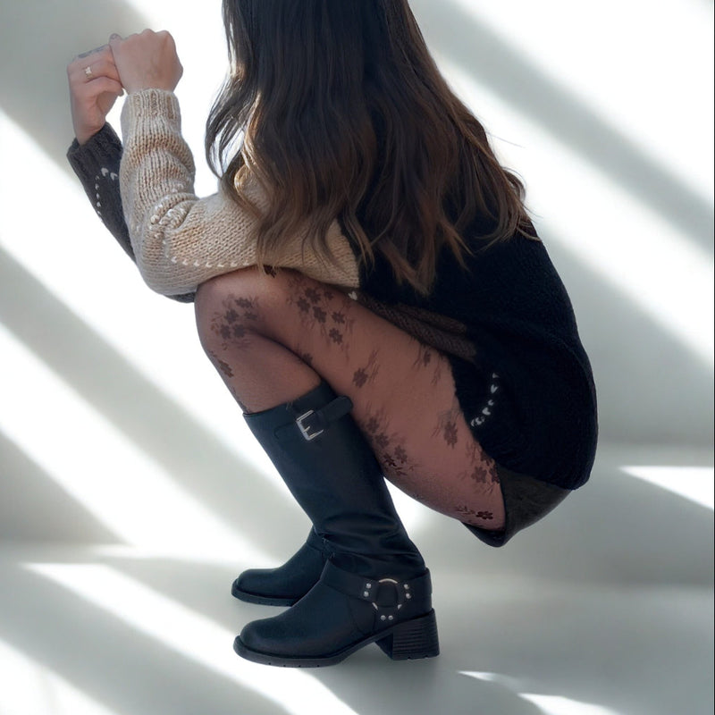 Person wearing black knee-high boots with buckles on a white floor with shadows.